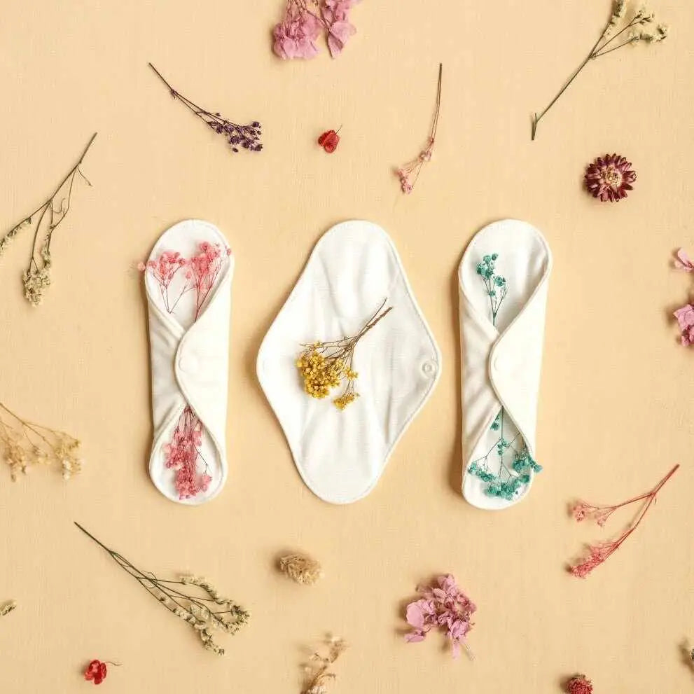 Tre assorbenti lavabili in tessuto bianco, due piegati e uno disteso con fiori secchi.