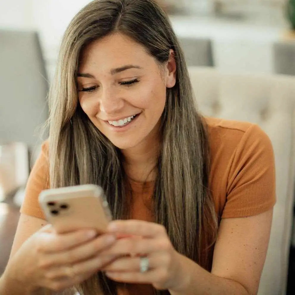 Donna sorridente con capelli castani lunghi indossa maglia rust a coste e usa smartphone su Yumibio