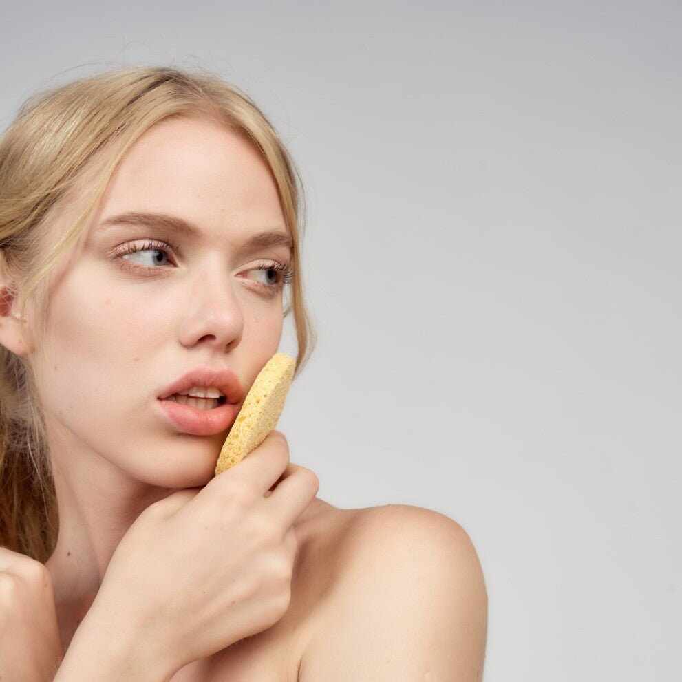 Giovane donna bionda con spugna gialla per viso sulla guancia, rimedio psoriasi