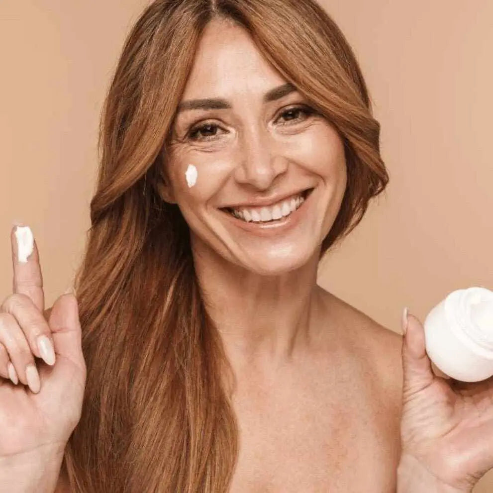 Donna sorridente con crema viso sul viso e dito, tiene vasetto antiage